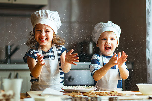 Two young children are dressed in chef&#039;s hats and aprons. They are laughing and clapping their hands. Food is flying in all directions.