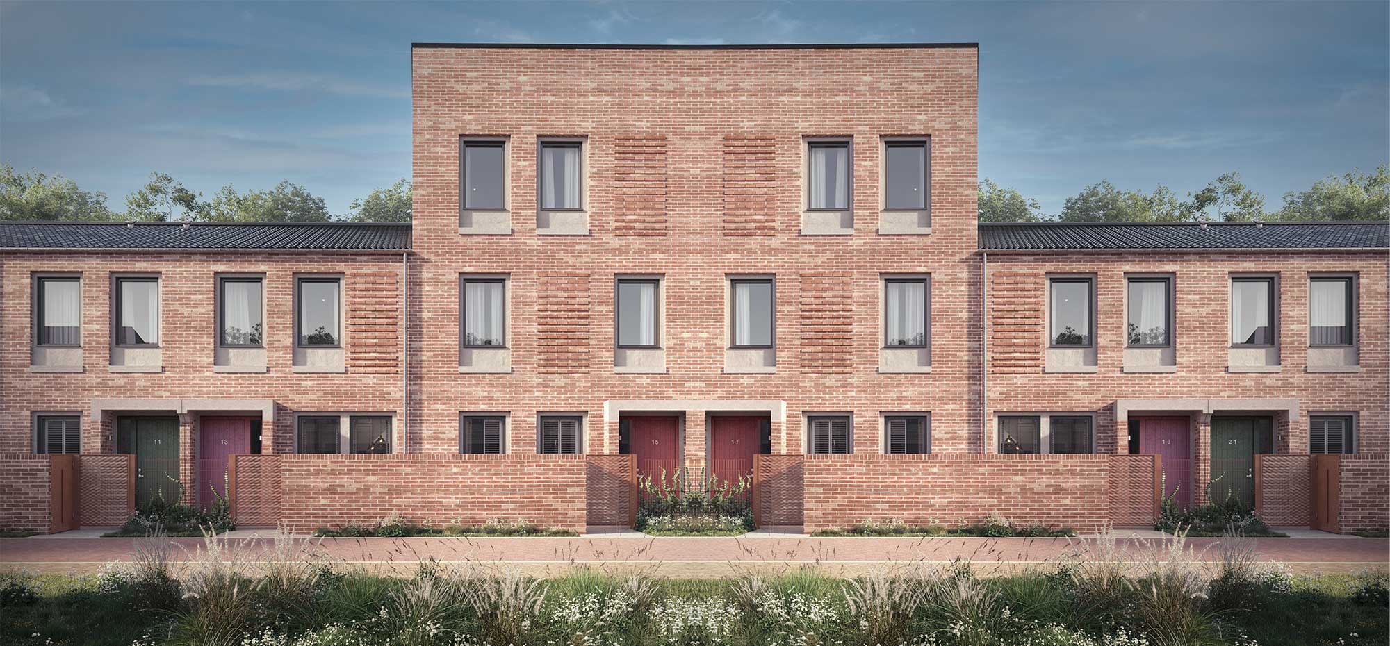Front view of a modern three-storey red brick terraced Passivhaus with large rectangular windows, some featuring shutters on the ground floor, and red and blue entrance doors, set behind a low brick wall and landscaped with grasses and wildflowers.